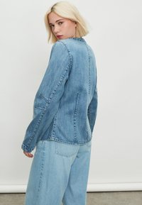 Világoskék farmerdzseki hosszú ujjakkal, testhez simuló szabással, varrási részletekkel és egyszerű gallérral. Világoskék farmernadrággal viselve.