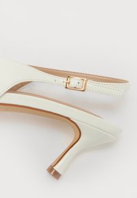 Zapato de tacón alto de cuero blanco con puntera afilada, tira delgada en el tobillo y detalle de hebilla en tono dorado. Textura suave con plantilla beige.