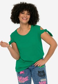 Groen T-shirt met korte mouwen en knoopaccenten op de schouders, ronde halslijn en een gladde textuur. Gecombineerd met blauwe jeans met kleurrijke tekst en ontwerpen.