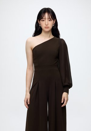 Vrouw met lang zwart haar die een donkerbruine one-shoulder jumpsuit draagt met één lange pofmouw, staand tegen een witte achtergrond.