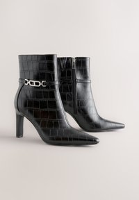 Black crocodile-patterned ankle boots with pointed toes, high heels, side zipper, and a decorative silver chain detail on the outer ankle.