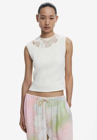 Top bianco a coste con un taglio in pizzo sul collo, indossato con pantaloni pastello a fantasia floreale con vita elastica.