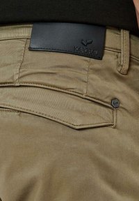 Pantalons kaki avec une texture lisse, présentant une étiquette en cuir noir au niveau de la taille et un petit accent hexagonal près de la poche.