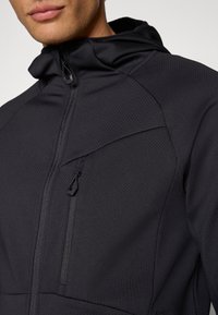 Giacca nera con cappuccio in tessuto strutturato, tasca frontale con zip e cordoncino regolabile al collo. Presenta un design aderente e dettagli delle cuciture.