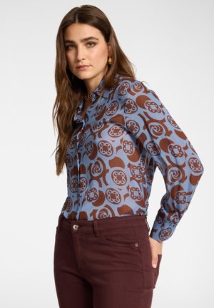 Jeune femme aux longs cheveux bruns portant un chemisier bleu avec des motifs circulaires marron et un pantalon rouge foncé, posant devant un fond blanc uni.