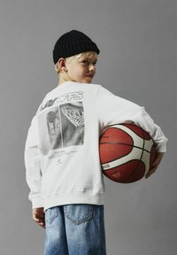 Witte sweatshirt met een basketbalafbeelding op de achterkant, met ribgebreide manchetten, gecombineerd met blauwe spijkerbroeken. Het kind houdt een basketbal vast.