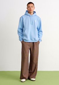 Lys blå bomull hettegenser med kengurulomme, kombinert med vide rutete bukser i brune, grønne og lilla nyanser. Hvite sneakers.