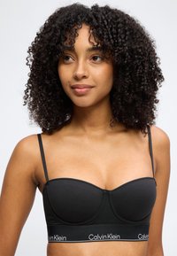 Ung kvinde med krøllet hår iført en sort Calvin Klein bralette, kigger svagt mod højre mod en ensartet lys baggrund.