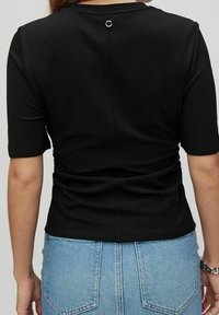 Haut noir à manches courtes avec une texture lisse, présentant un design froncé dans le dos et un accent circulaire argenté près de l'encolure.