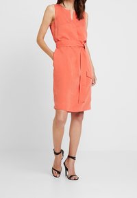 Robe corail sans manches avec un décolleté en V, ceinture à nouer à la taille, longueur au genou et poches. Se marie parfaitement avec des talons à bride à la cheville noirs.