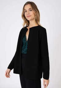 Gilet noir à ouverture devant avec des manches longues, en matière tricotée texturée et un col arrondi, superposé sur un haut à motifs vert foncé.