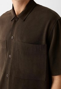 Ciemnobrązowa koszula z krótkim rękawem na guziki z dwiema kieszeniami na piersiach, noszona przez osobę na neutralnym tle.