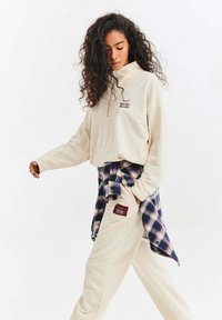 Béžová mikina s vysokým límcem a vyšitým logem, doplněná béžovými kalhotami s nášivkou. Vrstva s kostkovanou košilí uvázanou kolem pasu.