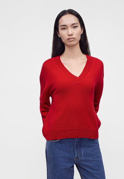 Femme aux longs cheveux foncés portant un pull rouge à col en V et un jean bleu, debout devant un fond clair uni.