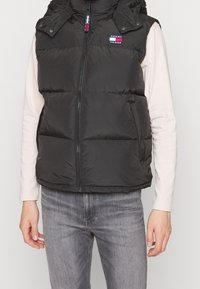 Schwarze Steppweste mit Reißverschluss und Kapuze. Verfügt über ein Logopatch auf der Brust, Seitentaschen und eine elegante, gesteppte Textur.