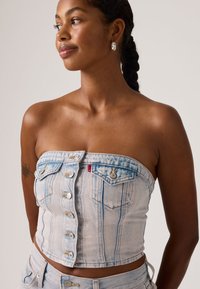 Γυναίκα με πλεξούδες, φορώντας αμάνικη ανοιχτό γαλάζιο ντένιμ μπλούζα με κουμπιά και ταιριαστό τζιν, κοιτάζει προς τα πλάγια με ασημένια κρίκους σκουλαρίκια.