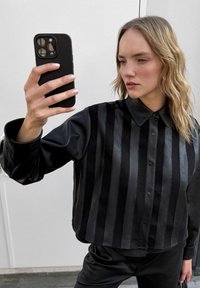 Czarna skórzana koszula z kołnierzykiem, z pionowymi paskami w różnych teksturach. Długie rękawy z zapięciem na guziki.