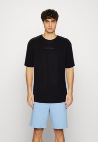 Sort bomulds T-shirt med korte ærmer og rund halsudskæring, med diskret branding på brystet. Kombineret med lyseblå shorts.