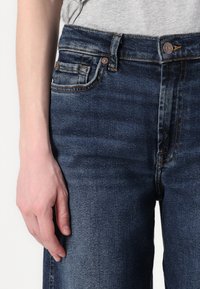 Dark blue denim jeans with a mid-rise waist, five-pocket design, and contrast stitching. Features a smooth texture and brass buttons.