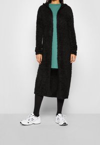 Schwarzer, flauschiger, langer Cardigan mit Kapuze, strukturiertem Stoff, langen Ärmeln, kombiniert mit einem grünen Shirt, schwarzen Leggings und weißen Sneakern.