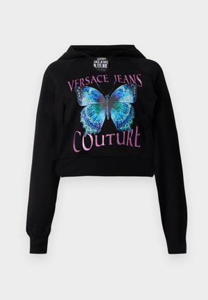 Melna īsā kapuce ar zila tauriņa grafiku un zaļiem akcentiem. Teksts "VERSACE JEANS COUTURE" metāliskā rozā krāsā. Mīksta tekstūra.