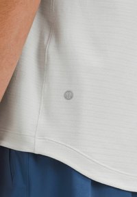 Weißes, strukturiertes Sportoberteil mit kleinem Logo, kombiniert mit blauen Shorts. Verfügt über sichtbare Nähte und eine lockere Passform am Saum.