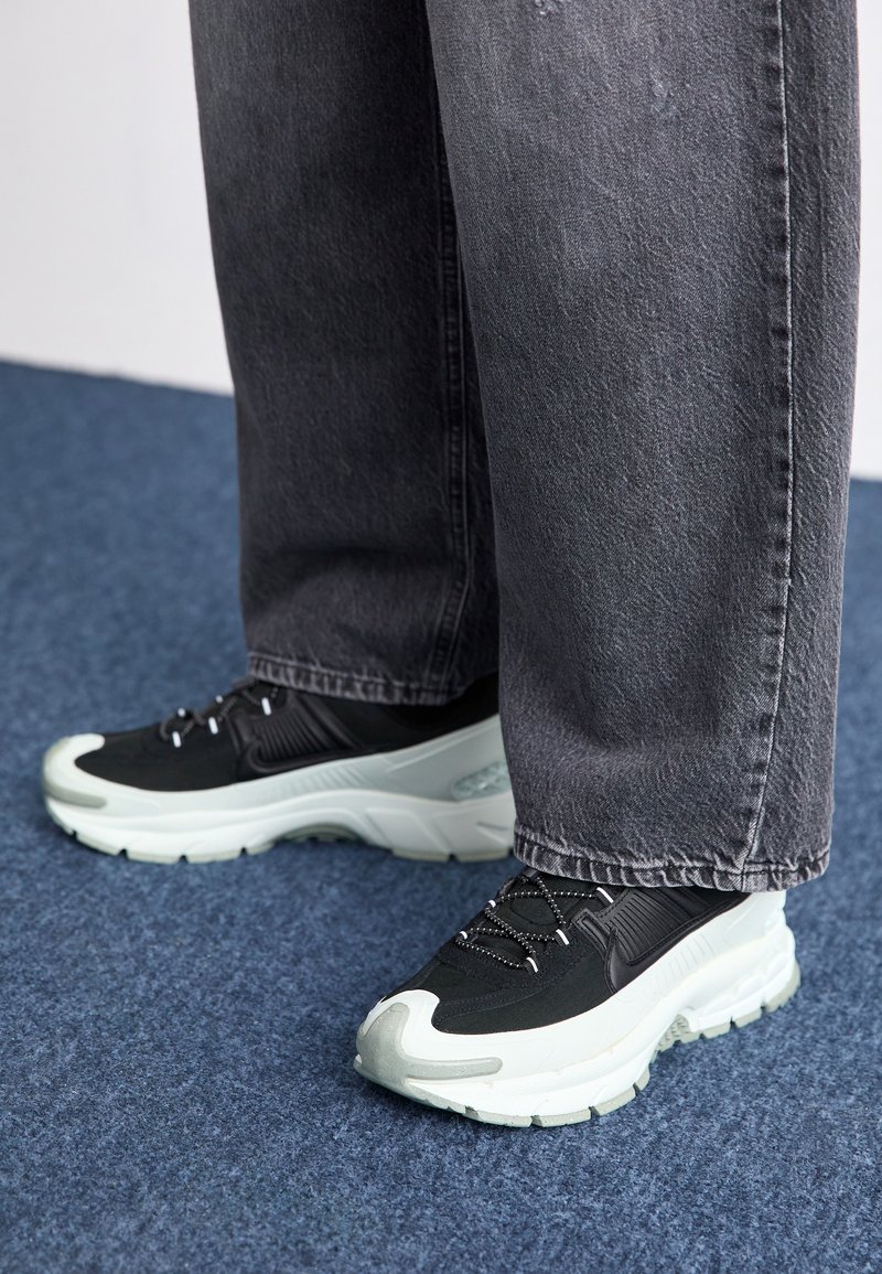 Czarne buty sportowe z fakturowaną cholewką, białą podeszwą środkową, czarnymi sznurówkami i gumową podeszwą, w połączeniu z szerokimi, szarymi dżinsami.