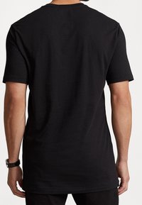 Muž stojící zády, oblečený v jednoduchém černém tričku s krátkým rukávem a černých kalhotách, na bílém pozadí.