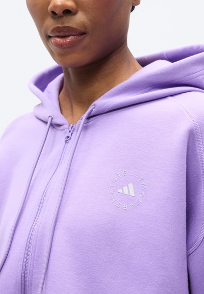 Ανοιχτό μοβ φούτερ με φερμουάρ, κατασκευασμένο από μαλακό ύφασμα, με κυκλικό λογότυπο στο στήθος και ρυθμιζόμενους κορδόνια στη κουκούλα.