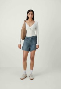 Valkoinen pitkähiha -paita kierretyllä yksityiskohdalla, yhdistettynä sinisiin farkkushortseihin, joissa on revitty helma, ja vaaleisiin nilkkakengiin. Ruskea olkalaukku korostaa asua.