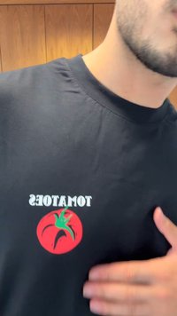 Camiseta negra con un gráfico de un tomate rojo con un tallo verde, y la palabra "TOMATES" en letras blancas en negrita sobre el diseño.