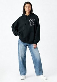 Sweat à capuche noir surdimensionné avec une poche frontale, présentant un dessin au trait blanc d'une colombe et le mot "Liberté". Associé à un jean large en denim clair.