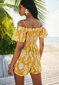 Mono amarillo con patrones florales blancos, mangas descubiertas y cuerpo fruncido. Tejido suave y estilo de pantalones cortos casuales.