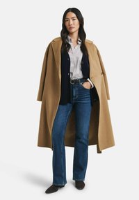 Camel-farbener langer Mantel, blaue Jeans, gemustertes Hemd, navyfarbener Cardigan, braune Stiefeletten. Sanfte Textur, taillierte Passform.