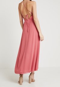 Maxiklänning i mjukt rosa tyg med ett korskorsdesign på ryggen, knytning i midjan, flödande kjol och ankellång längd. Neutrala högklackade sandaler.