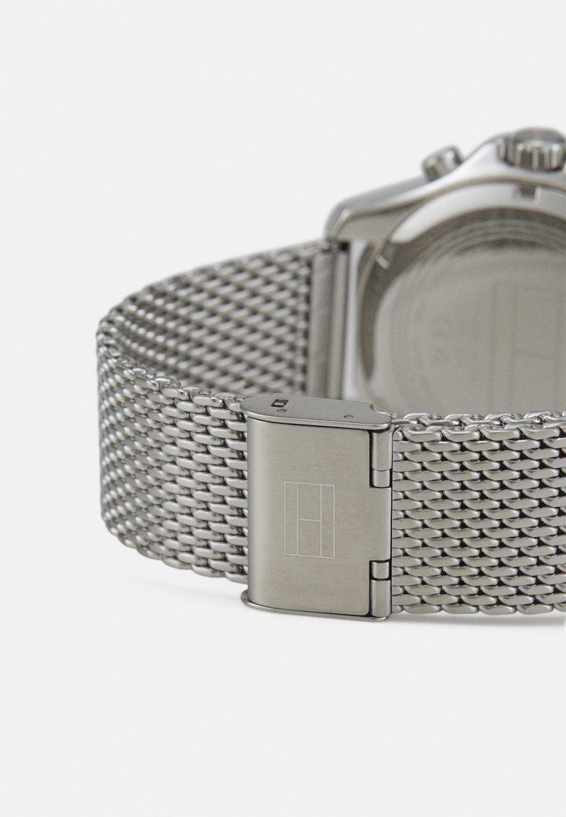 Correa de reloj de malla plateada con un cierre de acero pulido. Presenta una textura suave y una marca rectangular en el cierre. Esfera de reloj redonda.