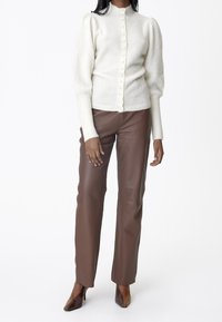 White knitted cardigan with puffed sleeves and a high collar, fastened with buttons, paired with brown leather trousers and pointed heels.