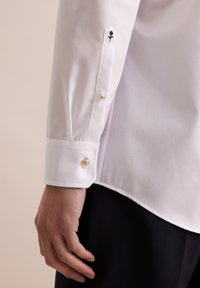 Camicia bianca a maniche lunghe con polsini abbottonati dotati di un bottone dorato e un fiore nero ricamato sulla manica. Tessuto dalla superficie liscia.