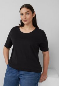 Junge Frau mit glattem braunen Haar trägt ein schwarzes Kurzarmshirt und blaue Jeans, steht mit einer Hand in der Tasche und der anderen auf einer weißen Fläche.