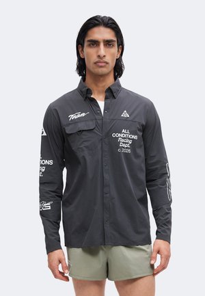 Homme aux cheveux noirs portant une chemise sombre à manches longues boutonnée avec du texte blanc et des logos, et un short vert clair, debout devant un fond uni.