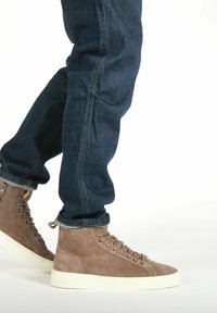 Brune suede højhælede sneakers med hvide gummisåler og sølvøjer, parret med mørke denimjeans, som er let op-rullet ved kante.