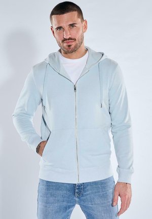 Man with beard wearing light blue zip-up hoodie and white t-shirt, standing with one hand in pocket against plain background.