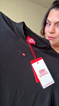 Chemise noire côtelée à manches longues avec un col monté. Présente un petit logo rouge sur la poitrine et des étiquettes rouges avec les détails du produit.