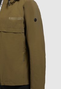 Giacca marrone con cappuccio, dotata di zip frontale, caratterizzata da una grande tasca, una patch con logo sul petto e un tessuto testurizzato. Design elegante e leggermente aderente.