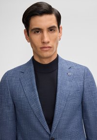 Blazer blu con texture e silhouette aderente, indossato sopra un dolcevita nero. Presenta dettagli discreti e un fermaglio con logo sulla patta.