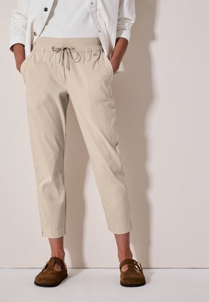Persoon die beige cropped broek met koord, wit shirt en bruine suède instapschoenen met gespen draagt, staand tegen een effen muur.