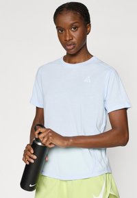 Αθλητική μπλούζα ανοιχτό μπλε με στρογγυλή λαιμόκοψη και κοντά μανίκια, με μικρό λευκό λογότυπο. Το άτομο κρατάει ένα μαύρο παγούρι νερού.