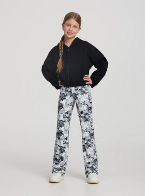 Dziewczyna z warkoczem w czarnej bluzie zapinanej na zamek, czarno-białych spodni wzorzystych i białych trampkach, stojąca z rękami na biodrach.