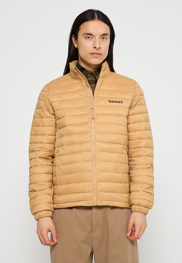 Giacca a vento imbottita color tan con cuciture orizzontali, colletto alto e chiusura con zip. Presenta un piccolo logo Timberland nero sul lato sinistro del petto.
