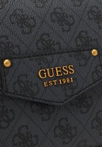 Struktureret sort materiale med præget mønster. Guld "GUESS" logo og detalje "EST 1981" på lommen. Funktionelt design med holdbart syning.